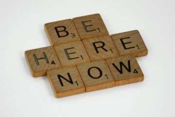 Inspirational text 'Be Here Now' created with wooden Scrabble tiles on white background.