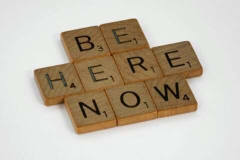 Inspirational text 'Be Here Now' created with wooden Scrabble tiles on white background.