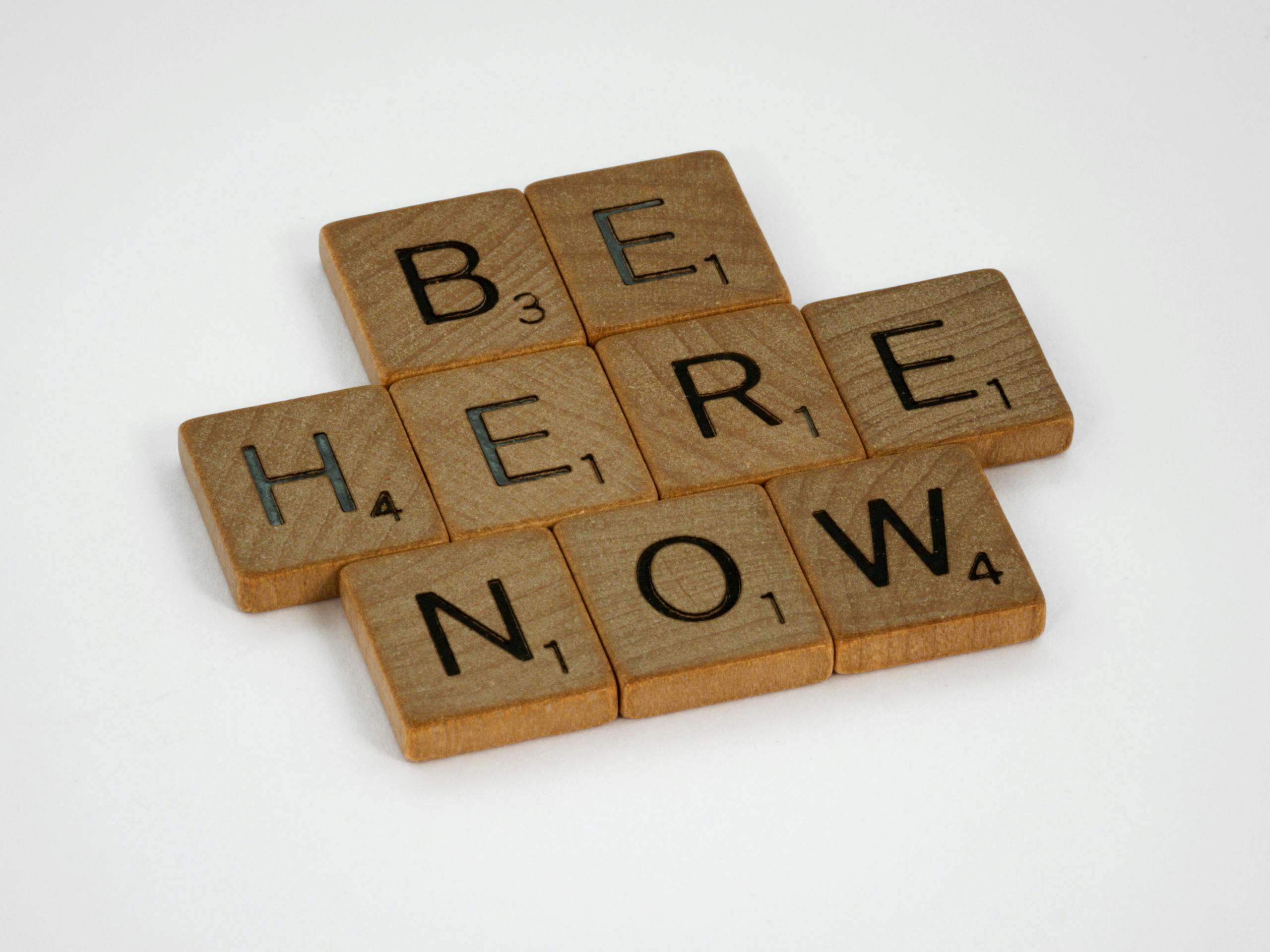 Inspirational text 'Be Here Now' created with wooden Scrabble tiles on white background.